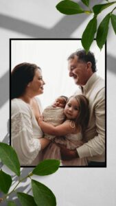 A family portrait documenting them welcoming a new baby. Mom and dad hold their toddler aged daughter and newborn daughter between them as they share a loving look. 