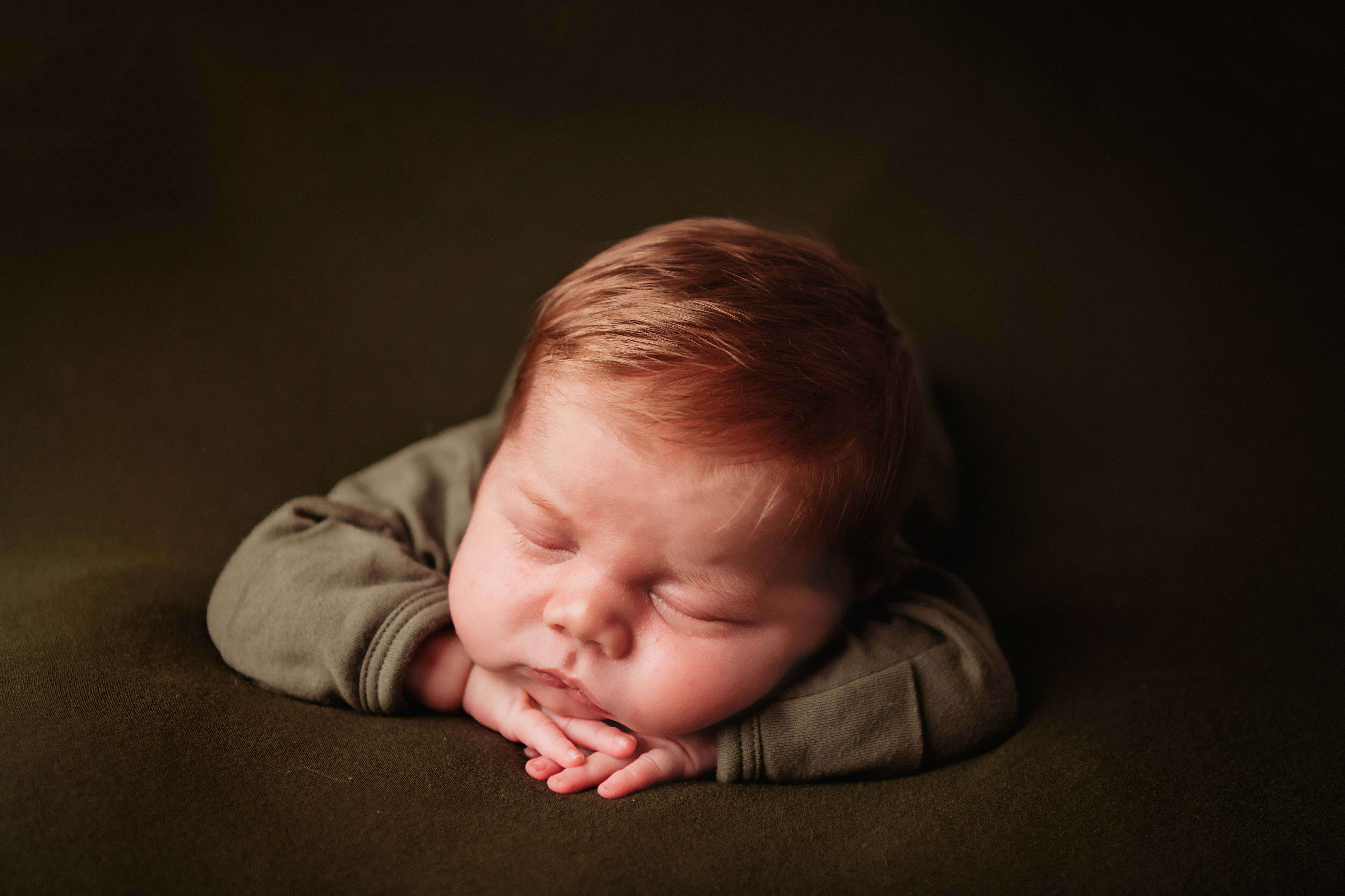 A red headed newborn boy, sleeping on his belly with his chin resting on his head. He is wearing green pajamas, and is resting on a matching green backdrop.